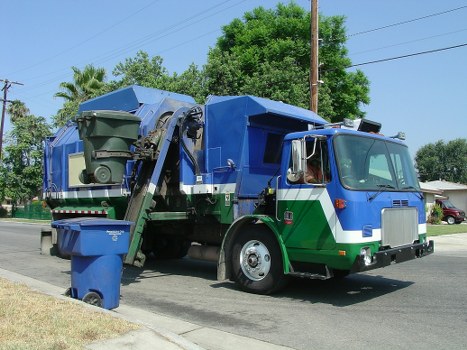 A large blue and green rubbish collection truck parked on a residential street with a clear blue sky. The truck has a spacious cab with a silver front grille and side mirrors, and a rear waste compaction area fitted with a hydraulic lifting mechanism. In the foreground, a black and green wheeled waste bin is positioned next to the truck, ready for collection. Surrounding the scene are trees, including a tall tree with dense green foliage behind the truck, and smaller trees and utility poles lining the road. The pavement and grassy verge are visible, indicating a typical suburban environment. The lighting is bright and natural, suggesting a sunny day. This image is relevant to professional rubbish removal services such as those offered by Office Clearance Putney, serving areas including Putney and nearby London neighborhoods.
