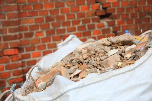 A large white polypropylene rubble sack filled with a mixture of construction debris including broken clay bricks, concrete fragments, and small wooden sticks, situated outdoors on a paved surface in front of a red brick wall. The rubble appears discarded after building work or renovation, with some dust and loose materials visible among the debris. The environment suggests a typical rubbish removal scene, reflecting services provided by Office Clearance Putney to clear waste from residential or commercial properties in the Putney area, with the debris container positioned for collection or disposal.
