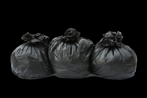 Three large black plastic rubbish bags are lined up horizontally, positioned on a flat surface with a black background. The bags are tightly knotted at the tops, with some slight creasing and folds in the plastic material. The bags appear to be filled with waste, giving them a rounded, bulging shape, and reflect a subtle sheen caused by the plastic texture. They are placed in close proximity to each other, with the middle bag slightly overlapping the other two. The setting is neutral, with a professional and observational tone suitable for a rubbish removal service, indicating disposal or collection of household or office waste, possibly in an outdoor or driveway environment typical for rubbish collection in Putney or nearby areas. The lighting highlights the bags’ contours and the textured surface of the plastic, emphasizing their fullness and portability for waste clearance tasks handled by companies like Office Clearance Putney.