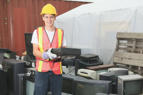 A young man wearing a yellow safety helmet and a high-visibility pink and yellow vest stands outdoors next to a collection of discarded electronic waste including old computer monitors, keyboards, and other electronic devices. He is smiling and holding a black plastic computer tower in his gloved hands, preparing to load or organize the waste materials. The background features a wooden fence, a white tarpaulin or temporary cover, and additional discarded items such as wooden pallets. The scene is set in an outdoor area that appears to be part of a waste collection or rubbish clearance operation, associated with professional rubbish removal services in Putney, close to the SW15 postcode district. The overall environment is well-lit, indicating daytime conditions, and the image emphasizes proper handling of electronic waste for environmentally responsible disposal.