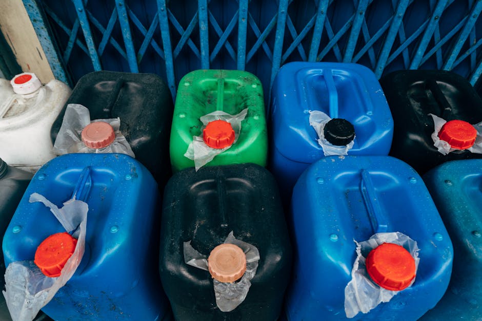 A large outdoor storage area filled with numerous plastic barrels arranged in overlapping rows. The barrels are primarily blue, with a handful of white containers featuring bright orange lids. The lid