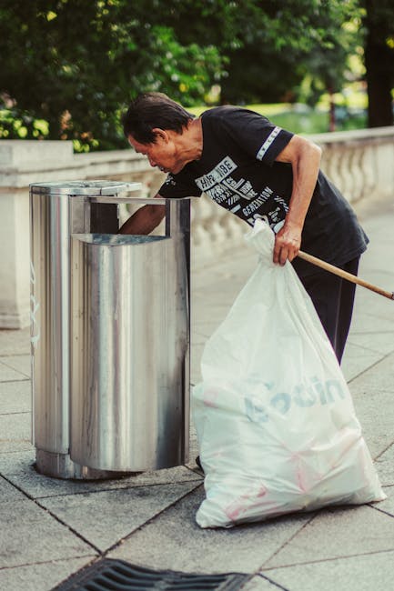 Two black plastic rubbish bags, filled and loosely tied, are placed on a paved sidewalk in front of a black metal fence with vertical bars. The bags appear to contain household or garden waste, with s