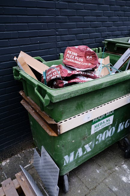 A large green plastic skip filled with several red and black bags of builder's waste, positioned outdoors against a dark brick wall. The bags are labeled with product information and appear to contain