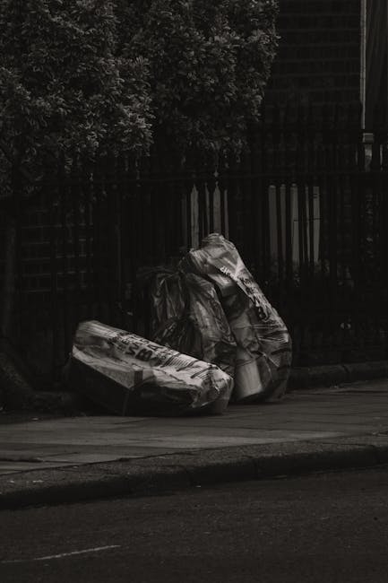 A large green plastic skip filled with several red and black bags of builder's waste, positioned outdoors against a dark brick wall. The bags are labeled with product information and appear to contain