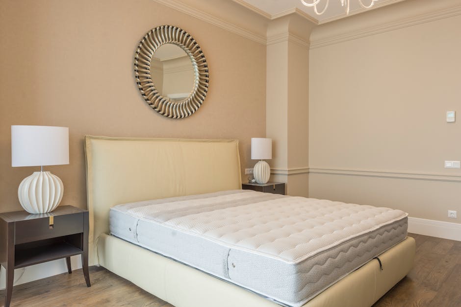 A minimalist bedroom interior with white walls and a light wooden floor. The room features a large double bed with a plain white mattress and a modern wooden headboard. On either side of the bed are s