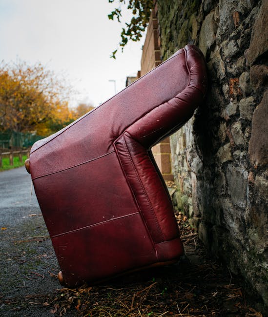 A beige upholstered armchair with a plush, velvety texture, placed outdoors against a weathered concrete wall with peeling paint and faded graffiti. The armchair has a high, rounded backrest with vert