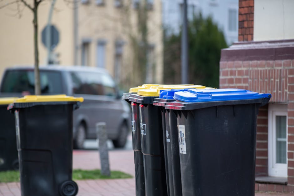 A row of three large black wheelie bins with closed yellow, blue, and black lids, positioned on a paved sidewalk next to a brick building. The bins are made of durable plastic with smooth textures and