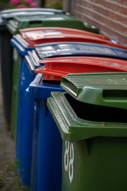 A row of three large black wheelie bins with closed yellow, blue, and black lids, positioned on a paved sidewalk next to a brick building. The bins are made of durable plastic with smooth textures and